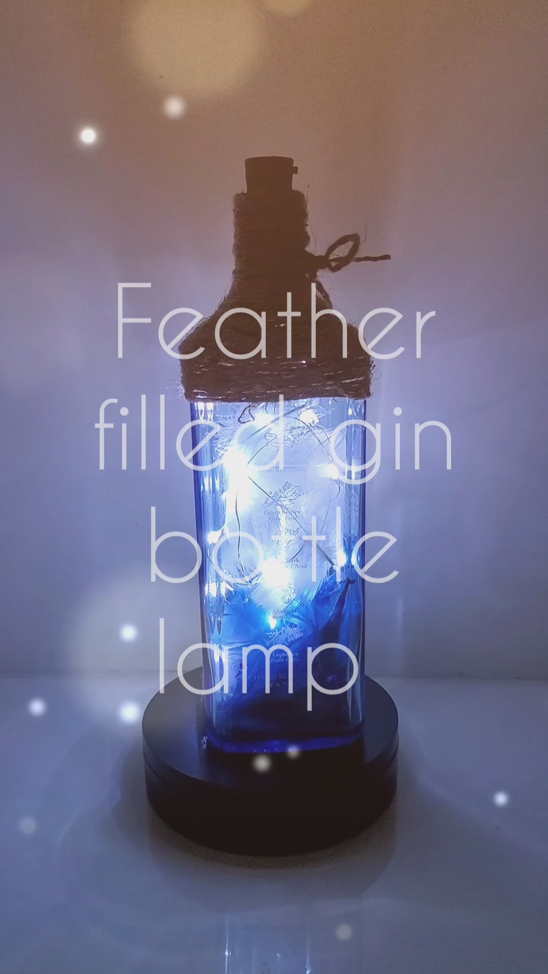 Rotating example of a Lit up blue glass gin bottle with jute twine detail around the top of the lamp, cork LED lights. Bottle is filled with blue and white feathers and blue coloured stones in the bottom.
