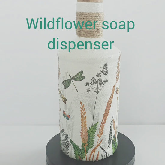 Gin bottle, painted white with wildflowers and dragonflies design. Silver pump head secured onto the cork of the bottle. Jute twine wrapped several times around the neck of the bottle. Rotating on a stand