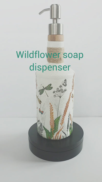 Gin bottle, painted white with wildflowers and dragonflies design. Silver pump head secured onto the cork of the bottle. Jute twine wrapped several times around the neck of the bottle. Rotating on a stand