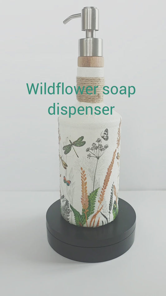 Gin bottle, painted white with wildflowers and dragonflies design. Silver pump head secured onto the cork of the bottle. Jute twine wrapped several times around the neck of the bottle. Rotating on a stand