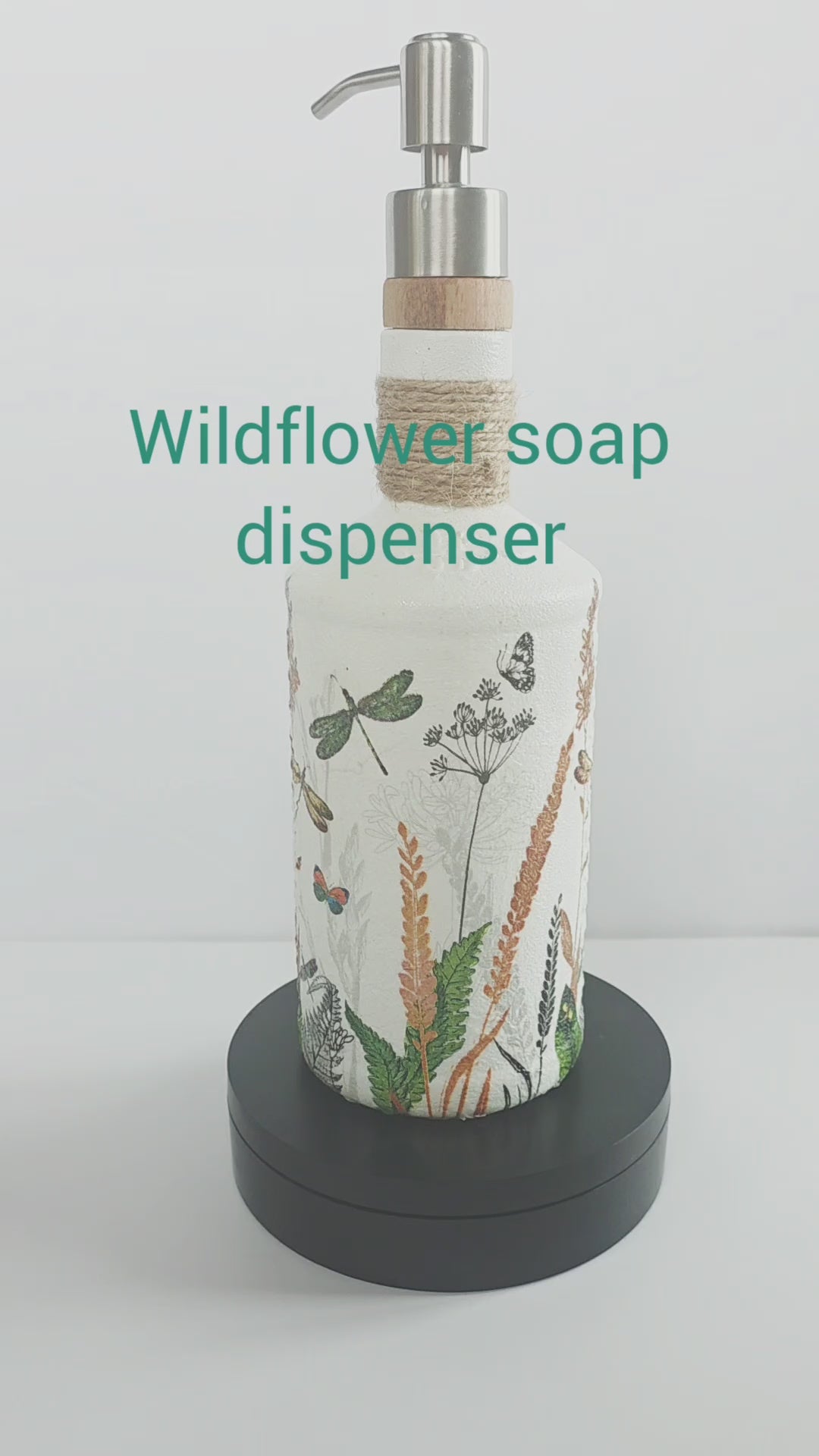 Gin bottle, painted white with wildflowers and dragonflies design. Silver pump head secured onto the cork of the bottle. Jute twine wrapped several times around the neck of the bottle. Rotating on a stand