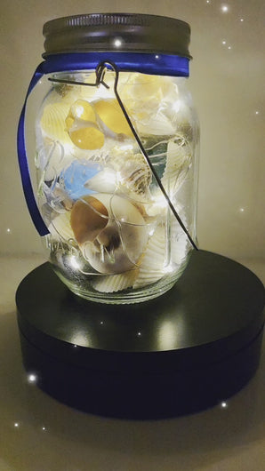 Jar filled with shells, pebbles and coloured glass stones. Lit up with lid and carry handle. Blue decorative ribbon