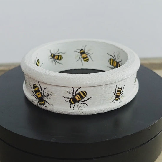 White wooden bangle with a ridged edge and bee design inside and out rotating on a black circular stand