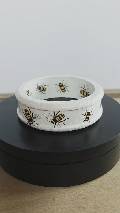 White wooden bangle with a ridged edge and bee design inside and out rotating on a black circular stand