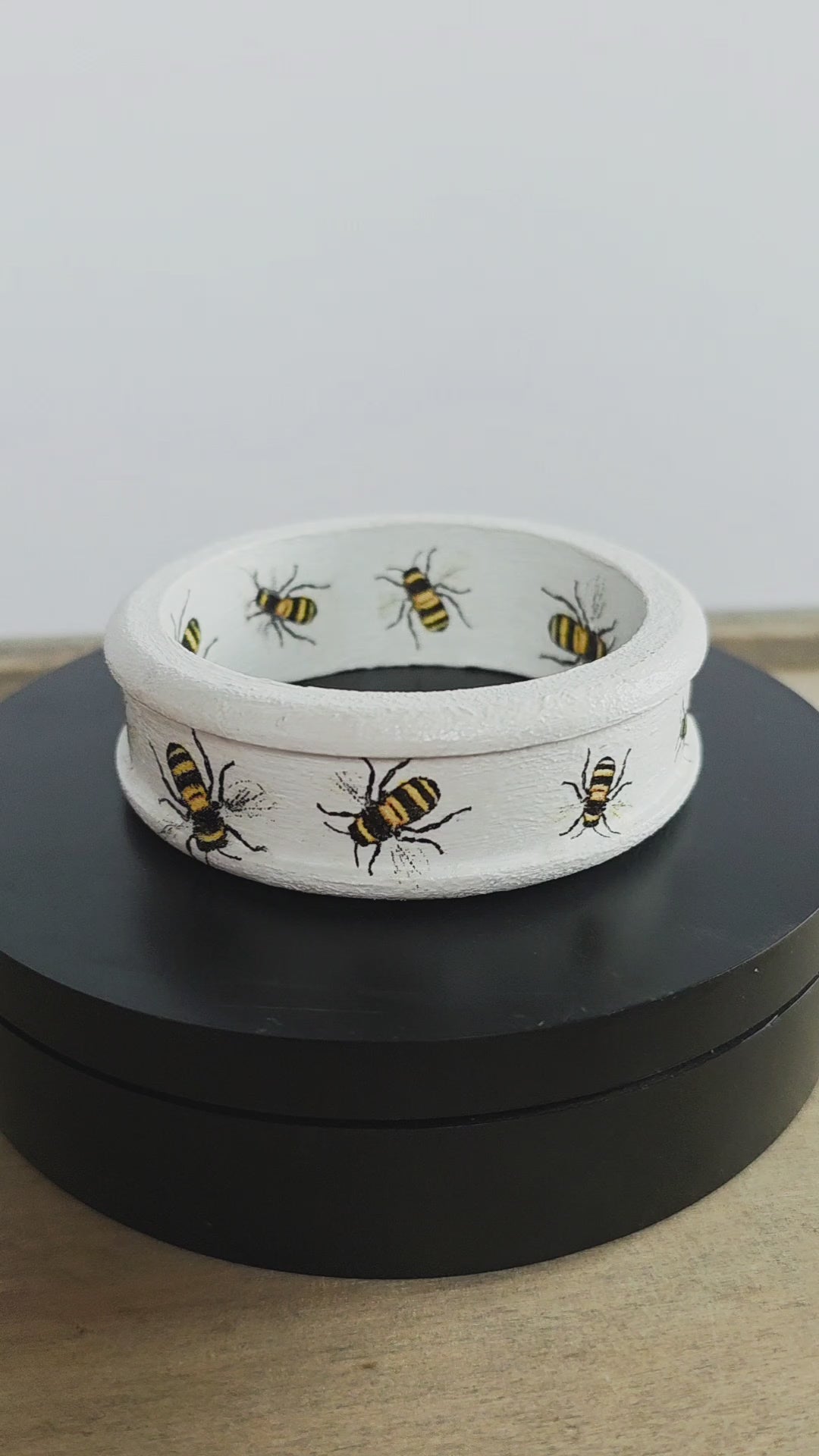 White wooden bangle with a ridged edge and bee design inside and out rotating on a black circular stand