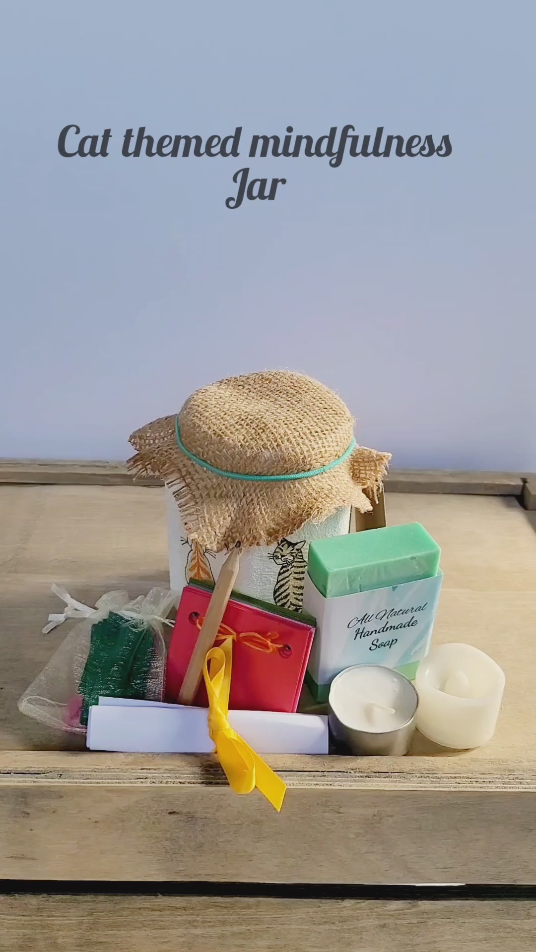 video of a white jar with a green bar of soap, tealight candle, paper scrapbook, brown pencil and small pale organza bag with green paper quotes, brown hessian lid