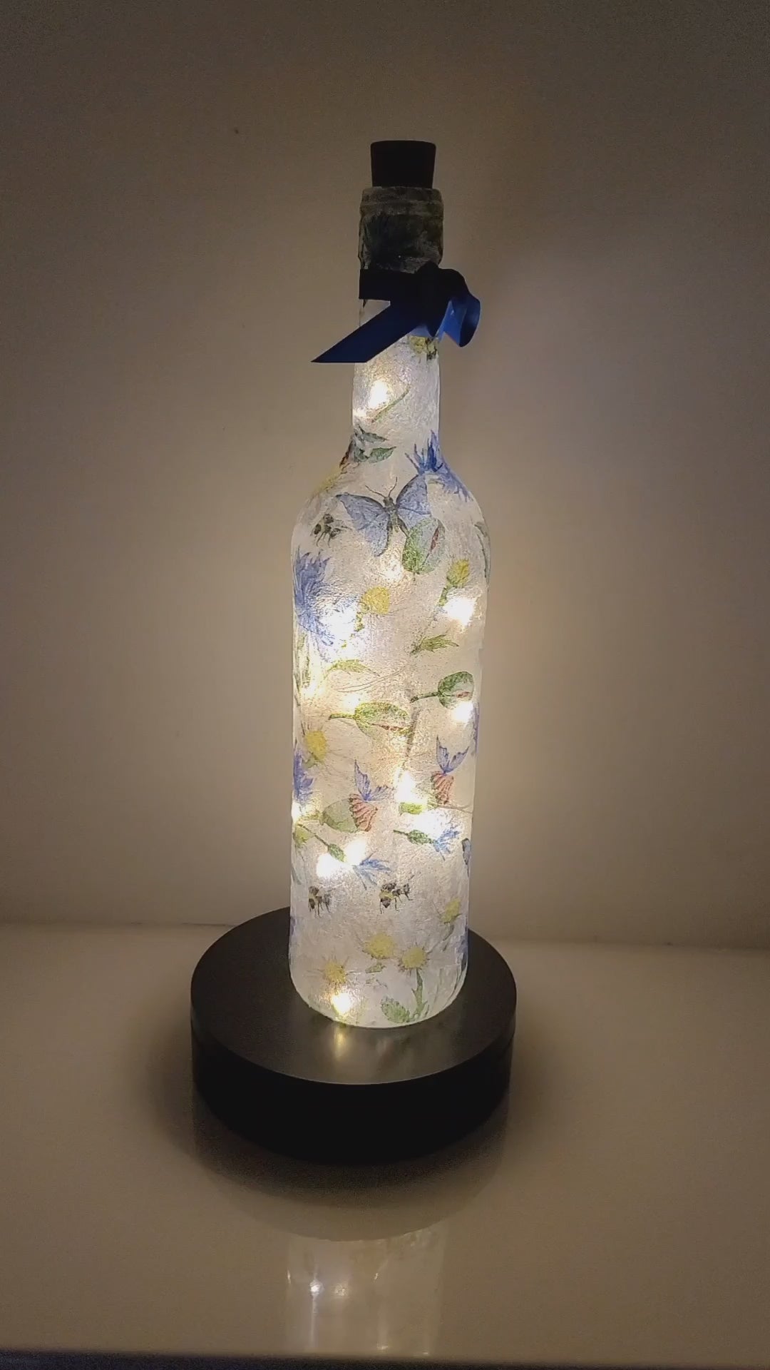 A rotating example of a A lit up wine bottle decorated with blue flowers, blue butterflies, acorns and bumble bee. Cork LED lights inside and blue ribbon around the neck of the bottle