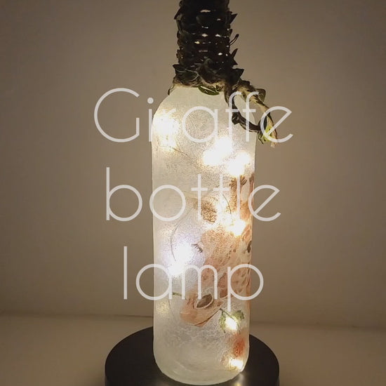 Rotating example of a Lit up wine bottle with single Giraffe detail and cork LED lights. Giraffe is holding a flower in its mouth. Jute twine with leaves is wrapped around the neck of the bottle