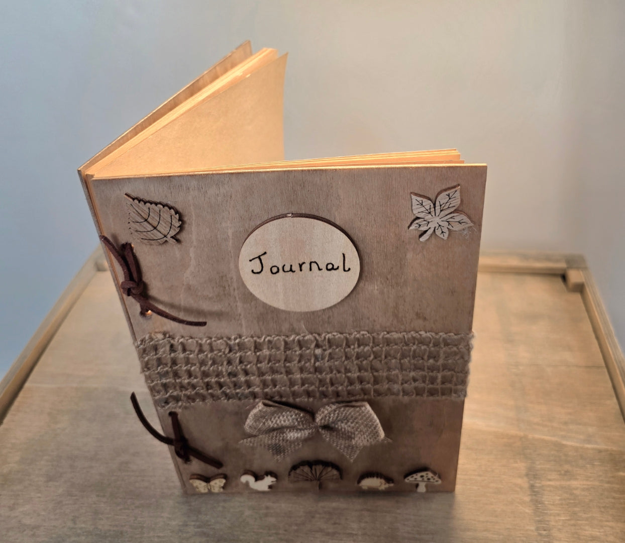 handmade rustic journal with wooden outercovers embellished with woodland creatures and wooden leaves. Burlap mesh decoration across the front, bound with brown suede, decorative hessian bow. Standing on its end and slightly open