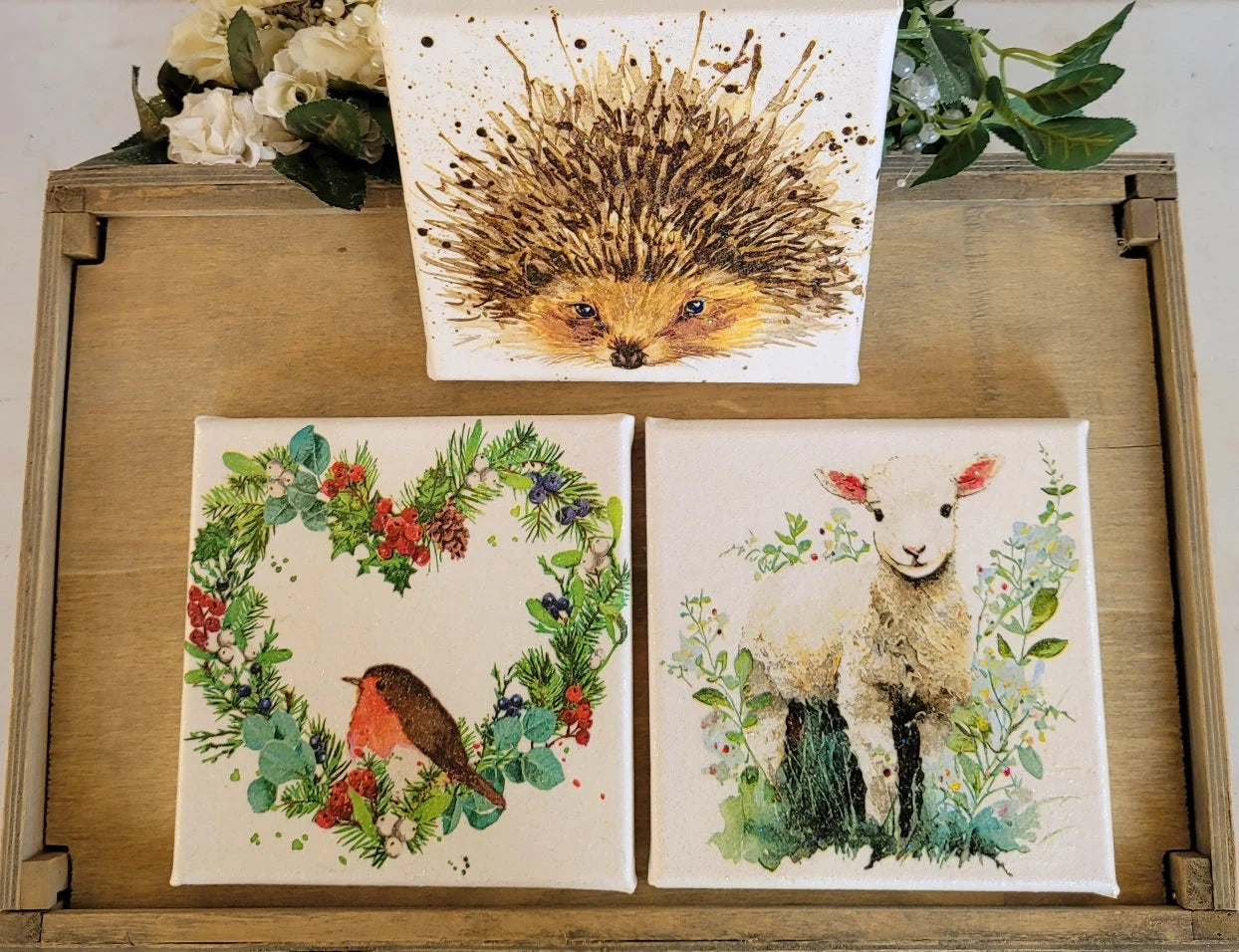 Three nature-themed prints featuring a hedgehog, robin, and lamb on a wooden surface with floral decorations.