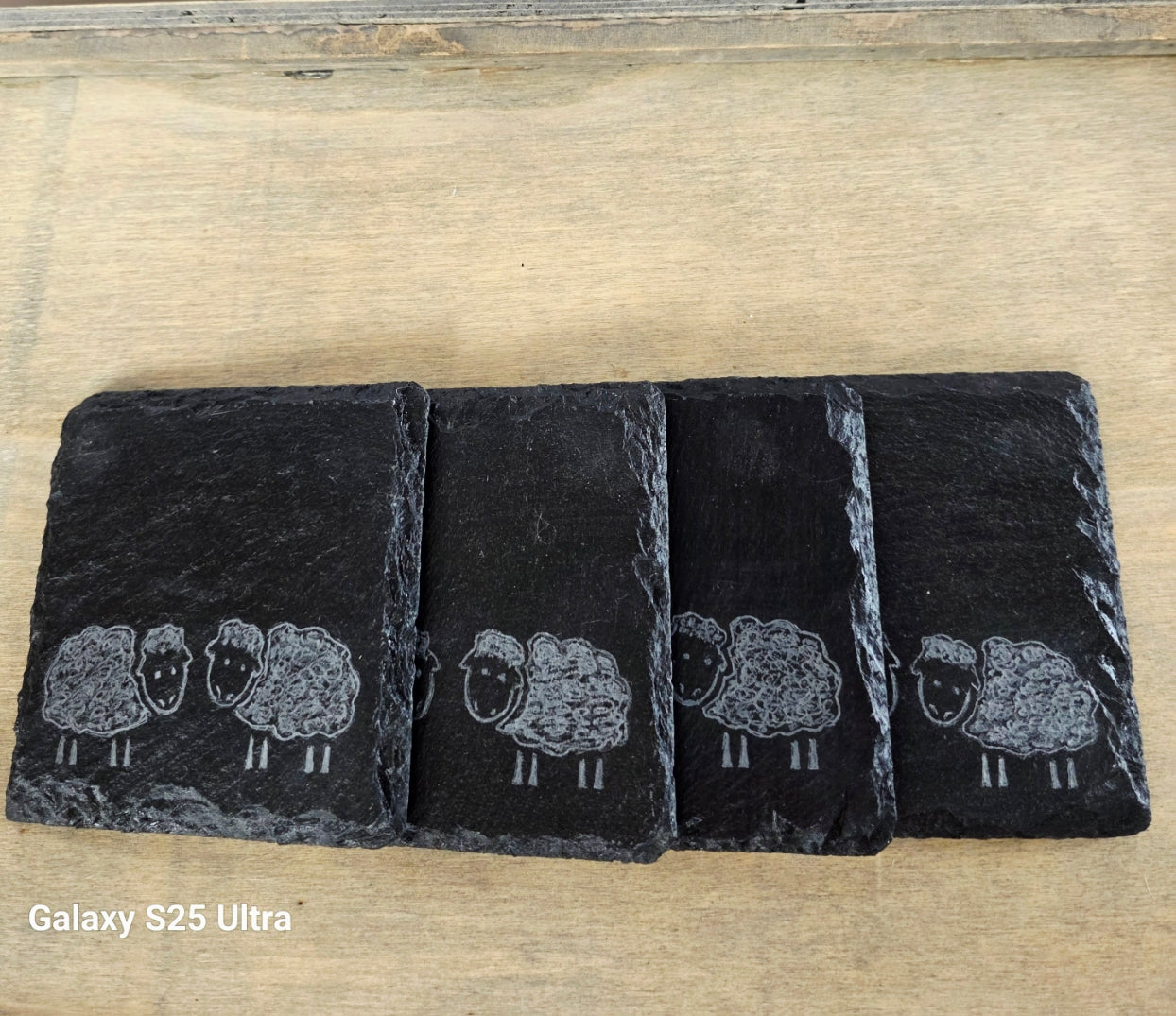 Four black slate coasters with white sheep illustrations on a wooden surface.