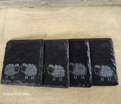 Four black slate coasters with white sheep illustrations on a wooden surface.