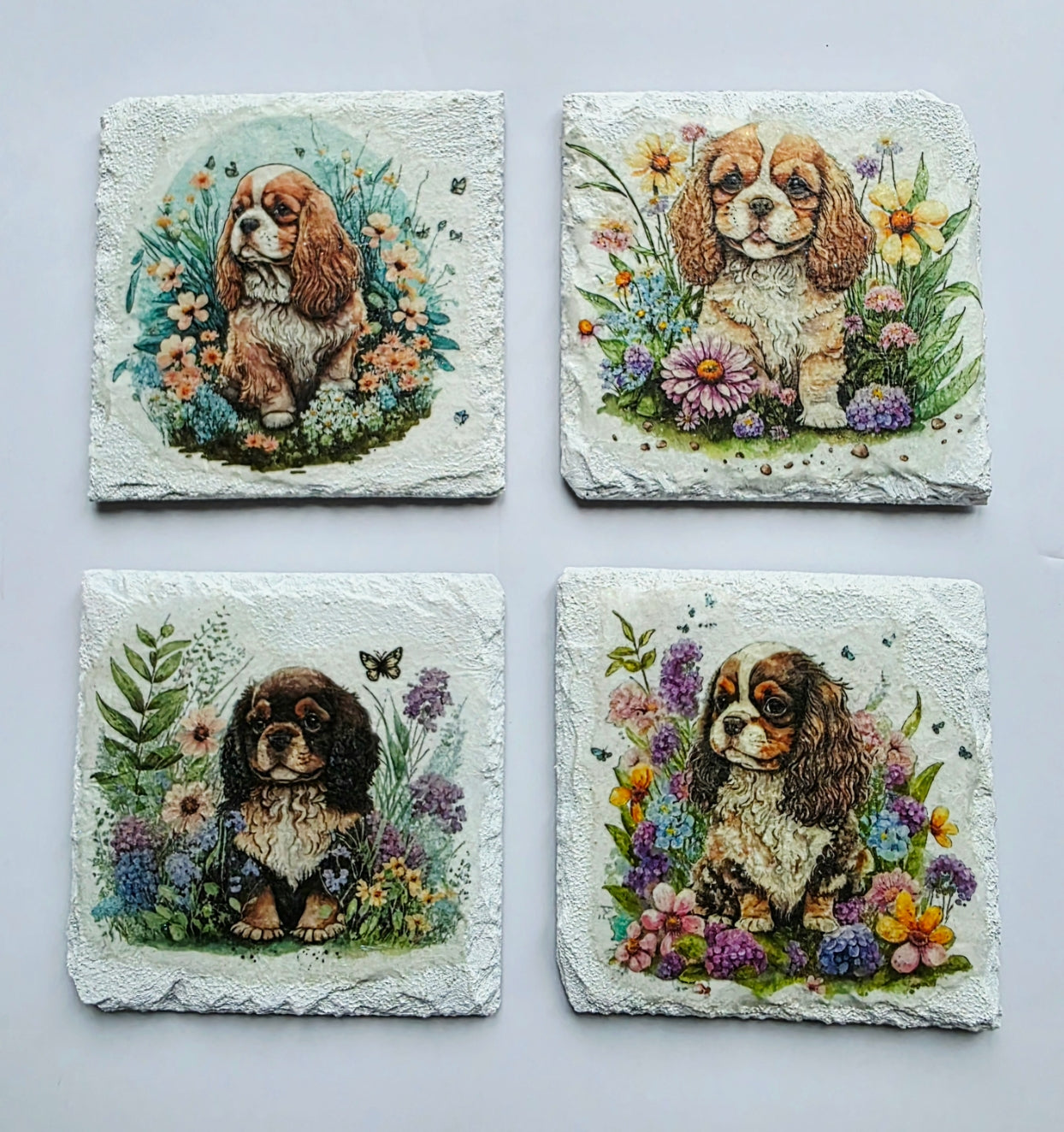 Four White slate coasters with four different paintings of a king charles spaniel puppy surrounded by flowers on a white background