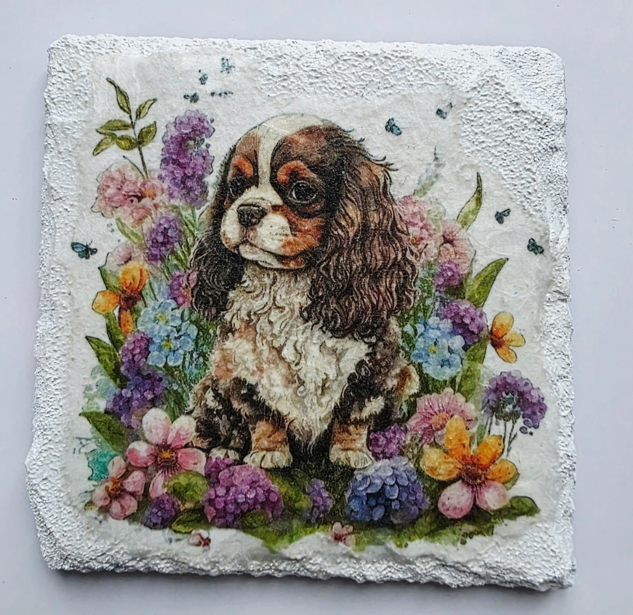 White slate coaster with a painting of a king charles spaniel puppy surrounded by flowers on a white background