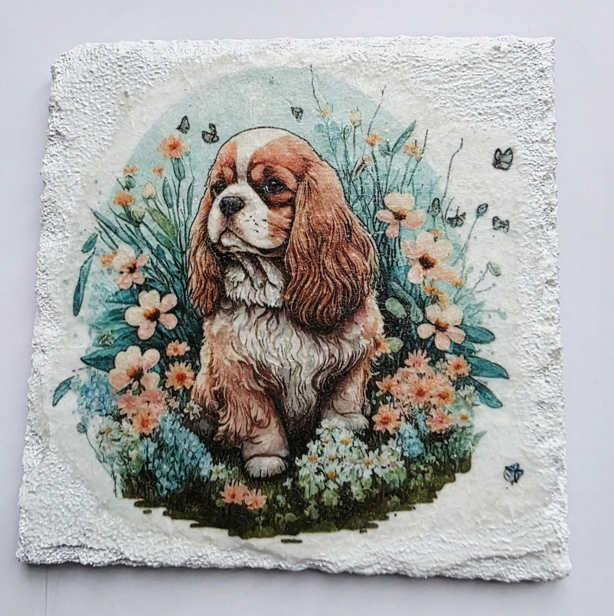 White slate coaster with a painting of a king charles spaniel puppy surrounded by flowers on a white background