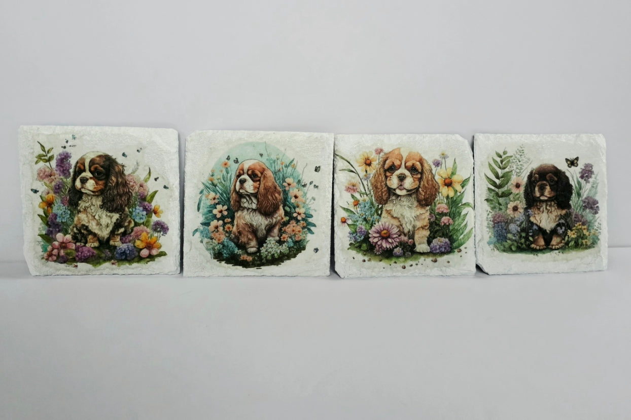 Four White slate coasters with four different paintings of a king charles spaniel puppy surrounded by flowers on a white background