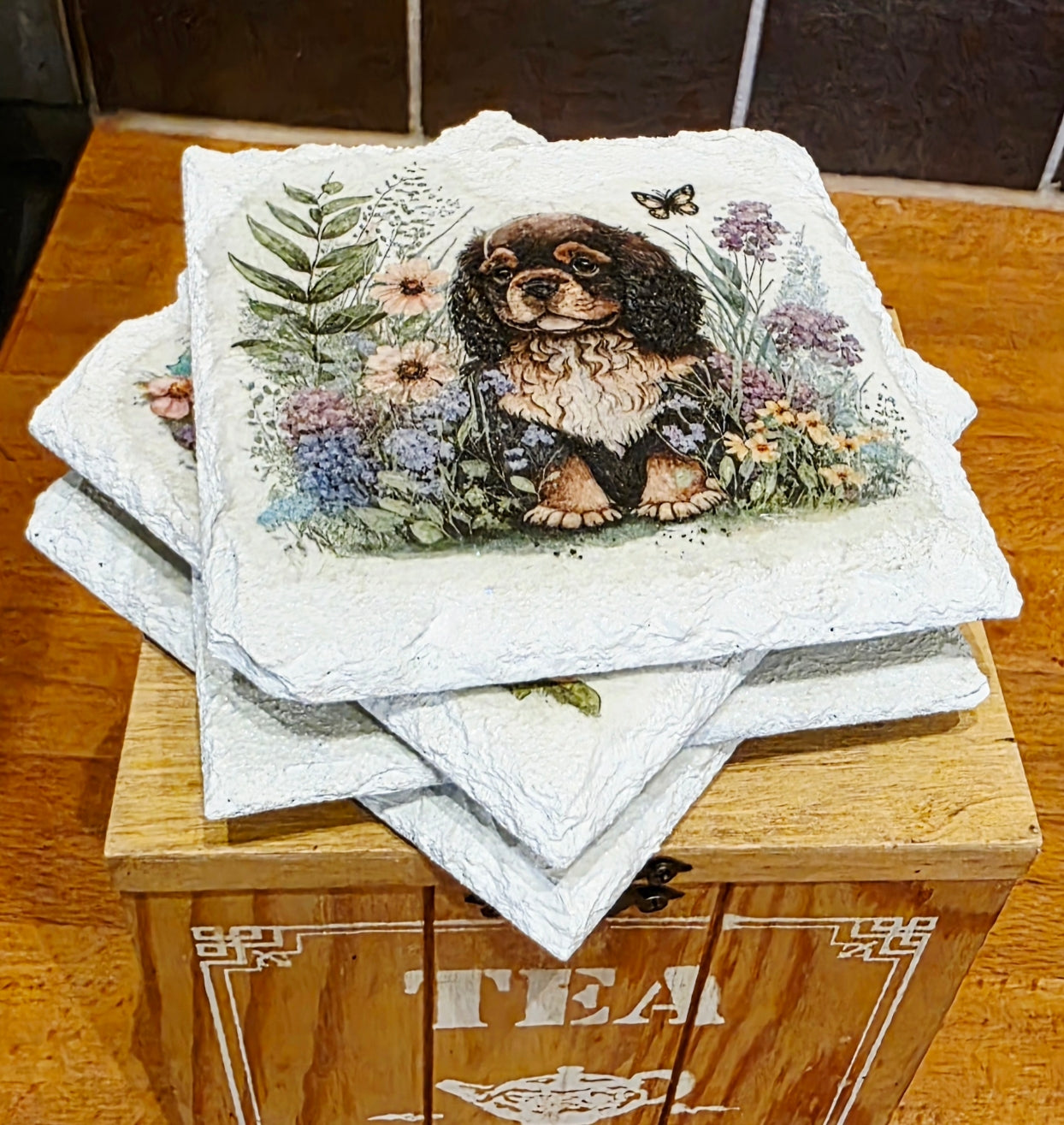 Stack of four white slate coasters with a spaniel dog and floral design on a wooden box labeled 'Tea'.