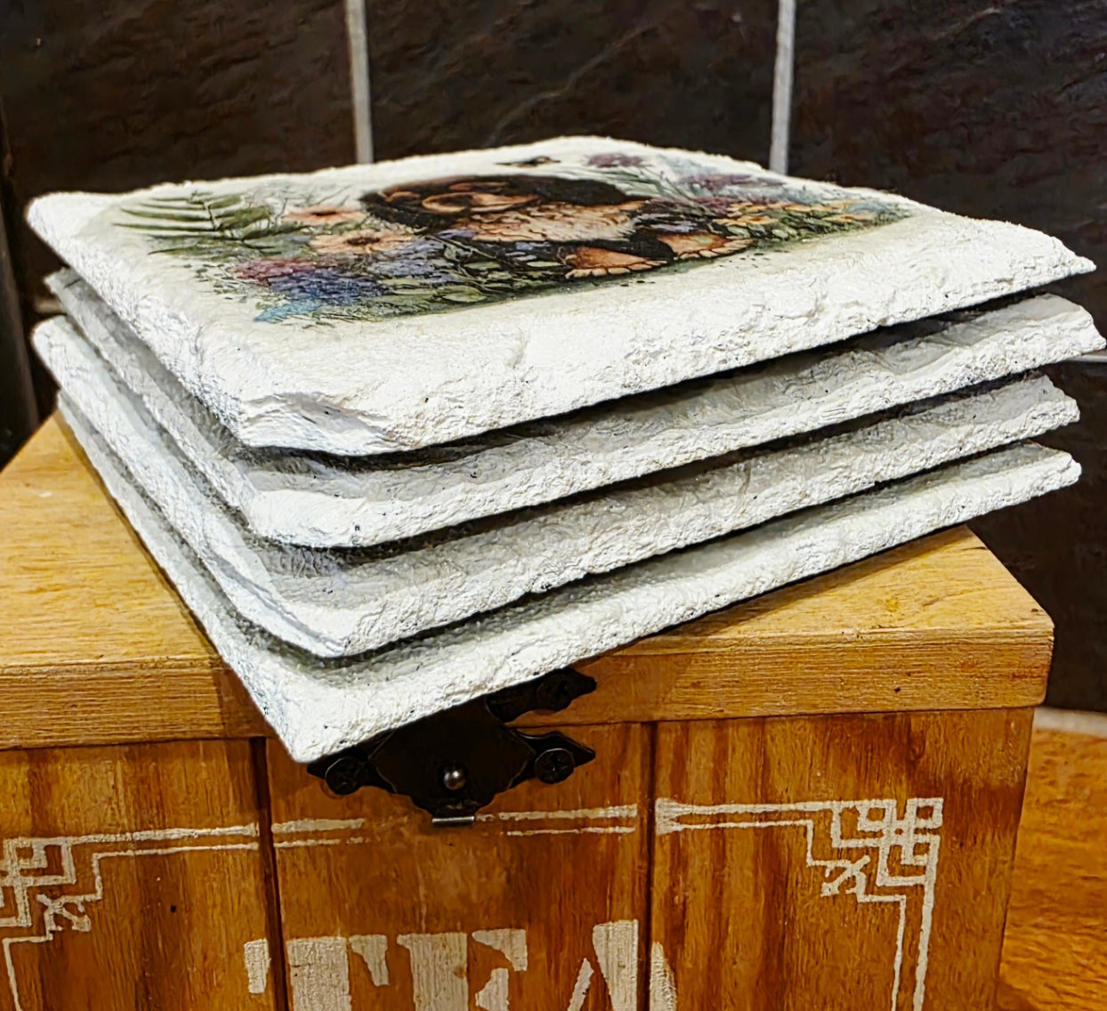 Stack of four white slate coasters with a spaniel dog and floral design on a wooden box labeled 'Tea'.