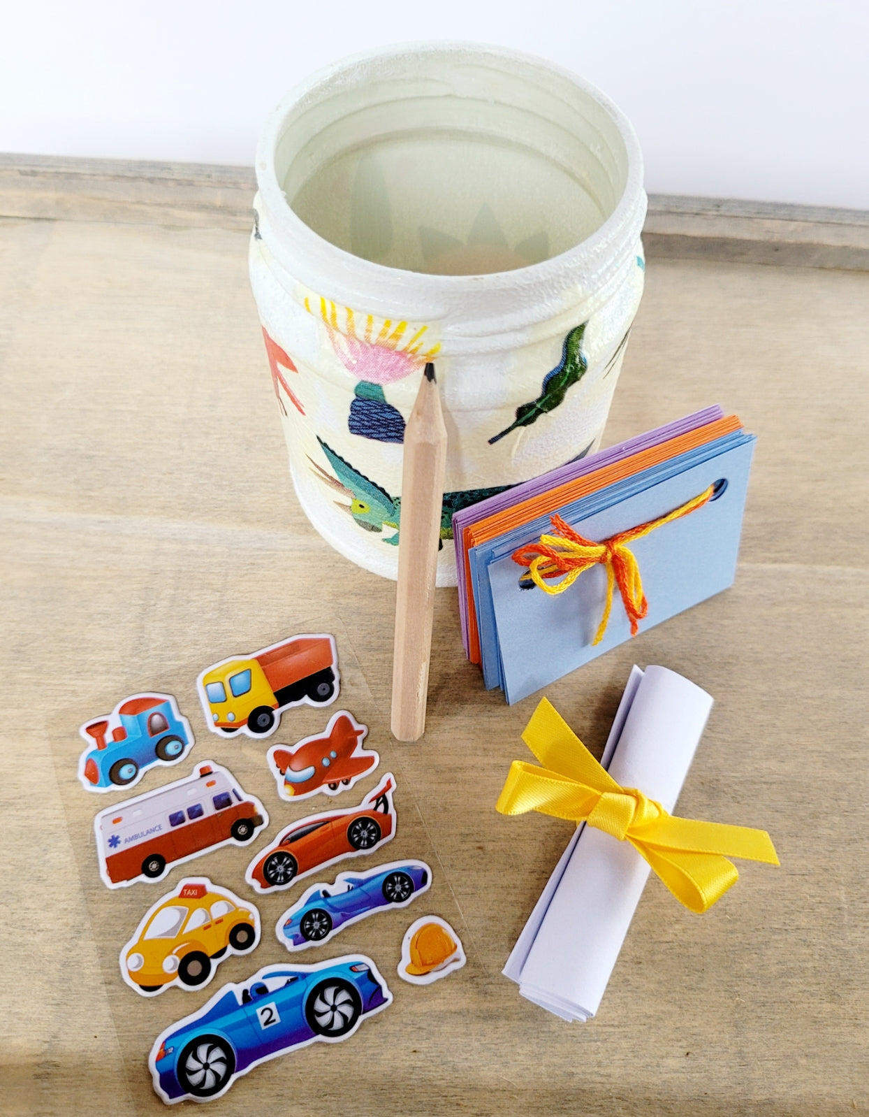 White painted jar with Dinosaur detail, small coloured scapbook with embroidery thread closure, a vehicle sticker sheet and short pencil. White rolled up note secured with yellow ribbon