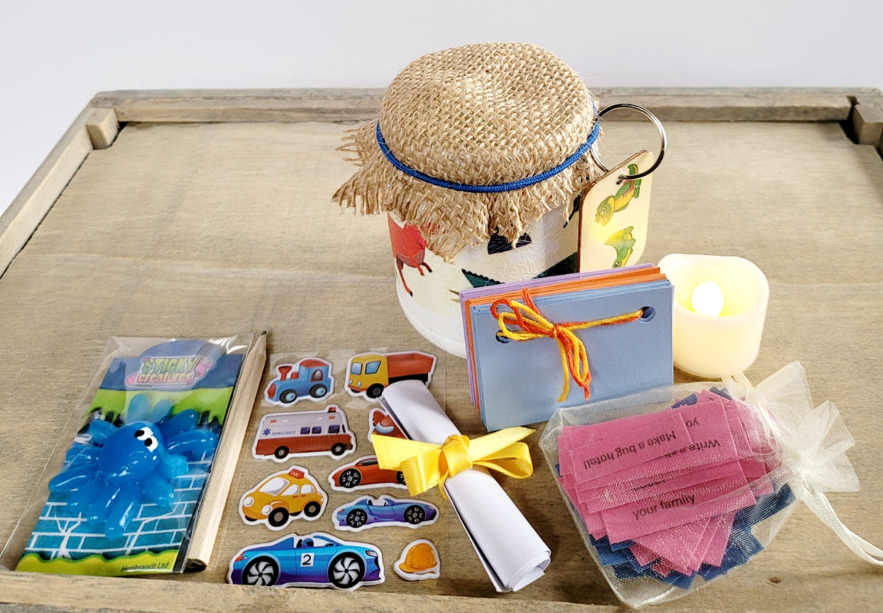 White painted jar with Dinosuar detail. Hessian lid secured with a blue band, sticker sheet, blue sticky toy, lit up flameless votive candle, white rolled up note secured with yellow ribbon, key ring attached to jar with a green Dinosaur on it. Coloured card scrapbook and short brown pencil, organza bag containing pink and blue quotes