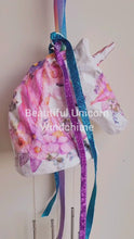 Load and play video in Gallery viewer, white paper mache Unicorn head with blue and purple sparkly ribbon. Pink flowers on the face of the unicorn, a pink unicorn on the side of the unicorn head. Silver chimes hanging below the Unicorn head
